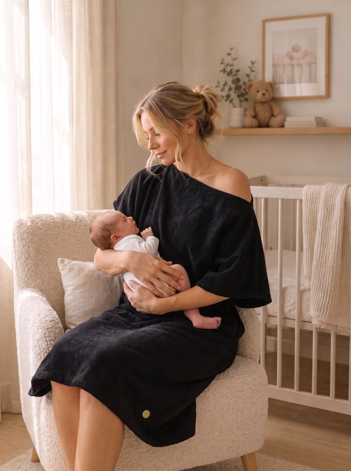 New mum in black Dry Dolly dress and poncho in nursery armchair with newborn
